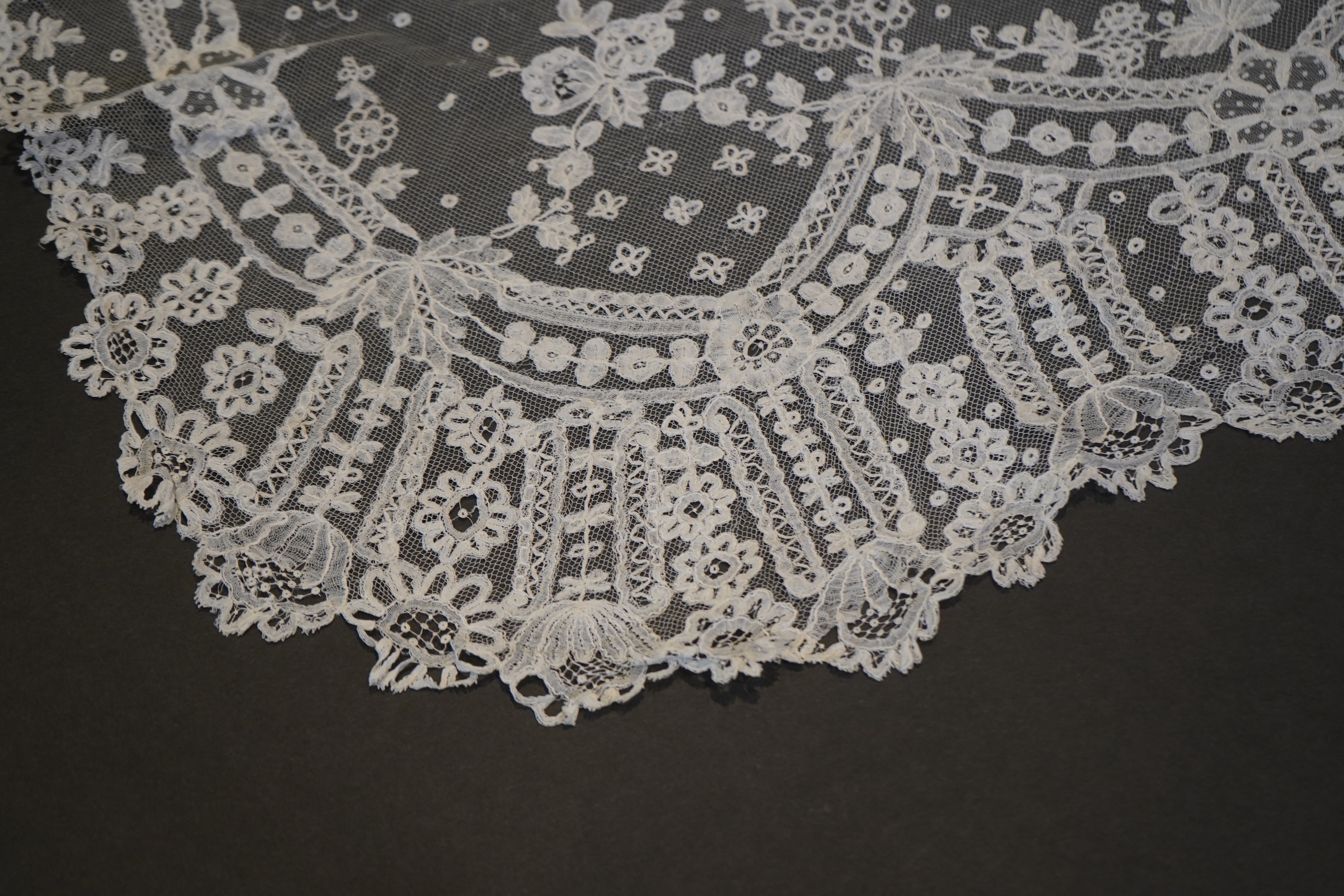 A deep elaborately designed flounce of mid 19th century Brussels bobbin appliquéd lace on net, with finished lace ends, worked with an intricate floral border below large floral cartouches framed in similar motifs and al